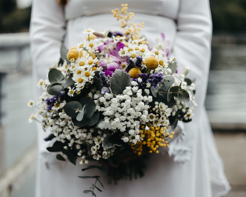 A person dressed in white, holding a large bouquet of fresh flowers arranged in a loose, natural style. The bouquet features white daisies, yellow billy balls, purple and white lavender, and silvery e
