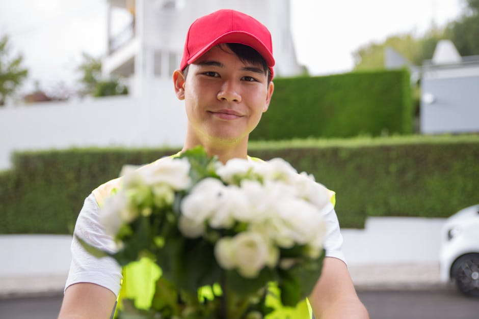 A delivery person with light skin, wearing a red cap and a high-visibility vest over a white t-shirt, smiling while holding a bouquet of fresh white roses and green foliage. The bouquet is slightly ou
